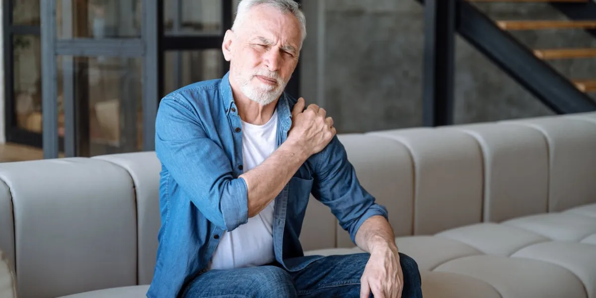 arthritis disease, problem with health senior man suffering from pain in shoulder, sitting on couch at home male touching, massaging inflammation zone