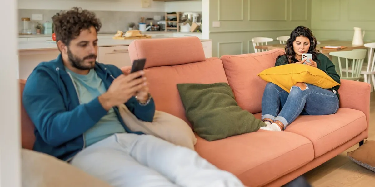 young married couple using smart phone ignoring each other addicted to the internet and social network - man and woman phubbing in the living room - family people loneliness and technology lifestyle concept
