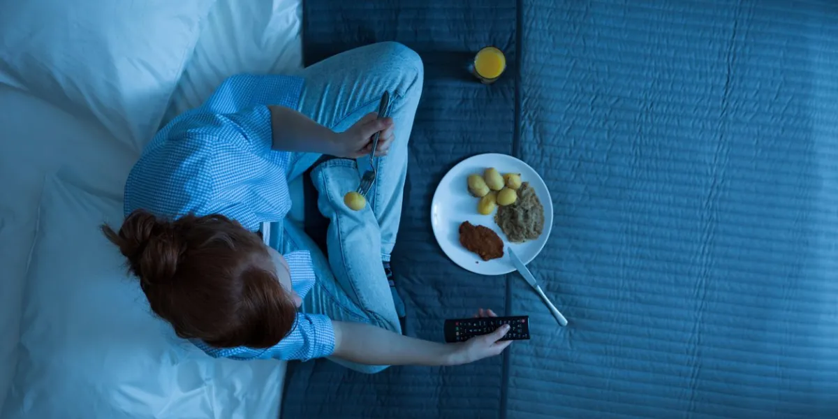 photo from the top of young girl sitting on a bed, eating dinner and watching television