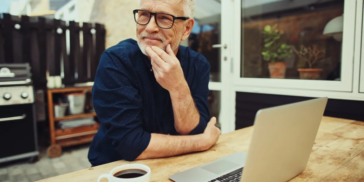 smiling senior man sitting at a table outside on his patio working online with a laptop and drinking a cup of coffee