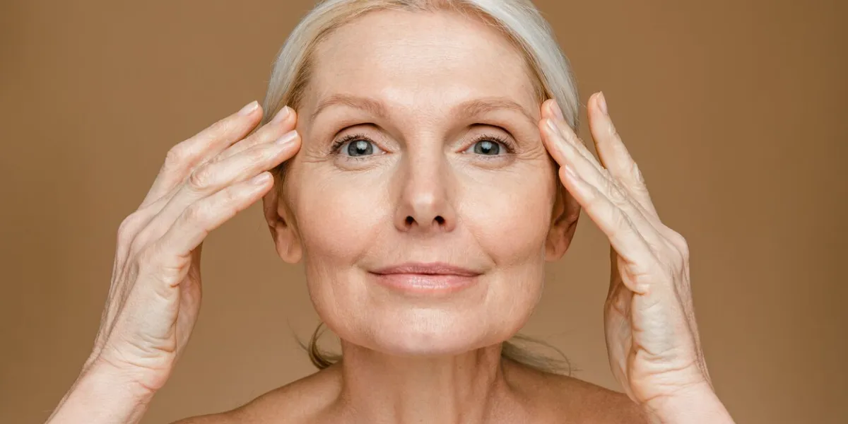 portrait of beautiful mature blonde woman touching her face, posing over beige brown studio background sensual senior lady with soft skin looking and smiling at camera