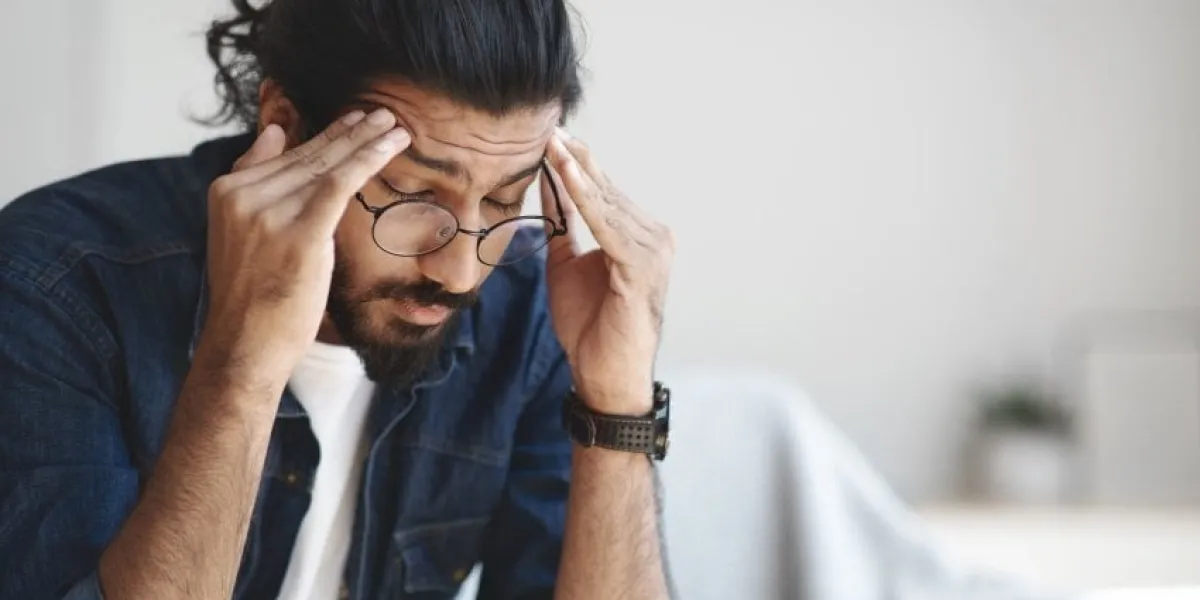 young indian guy freelancer suffering from headache after hard working day, sitting on couch at home, massaging head temples, having strong migraine, feeling tired, upset and stressed, copy space