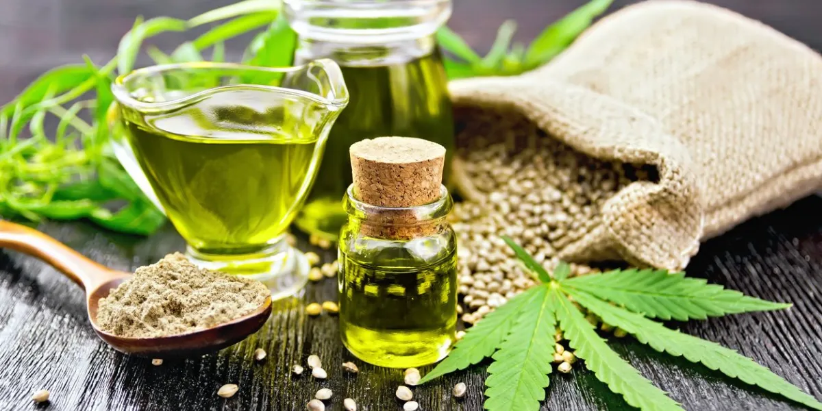 hemp oil in two glass jars and sauceboat with grain in the bag, leaves and stalks of cannabis, a spoon with flour on the background of wooden boards