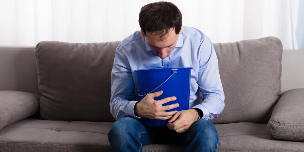 close-up of a man sitting on sofa vomiting at blue plastic bucket at home