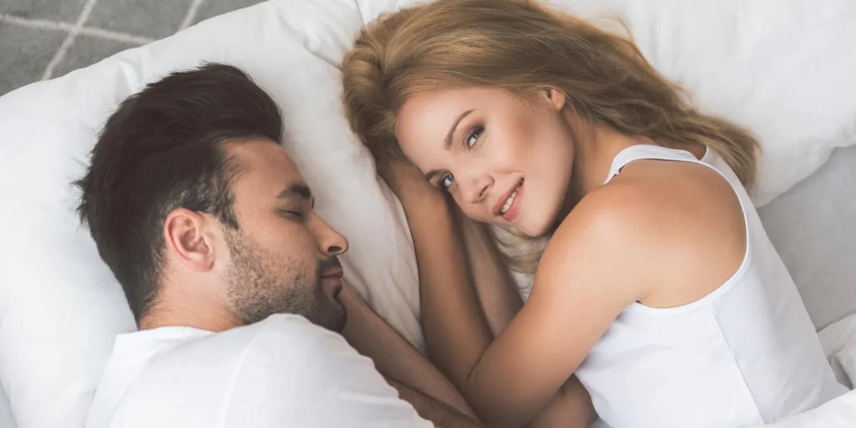 top view portrait of joyful girl relaxing on bed near her sleeping lover she is looking at camera with happiness and smiling
