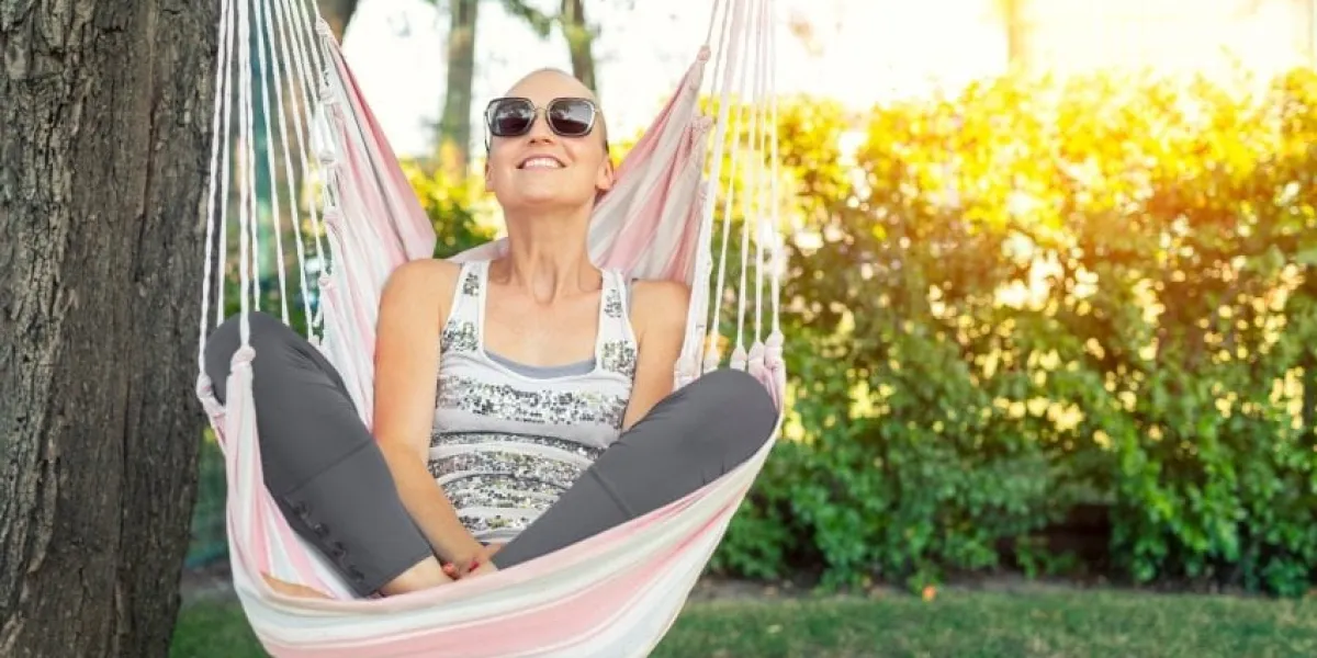 young adult happy short haired shaved bald attractive woman chilling enjoy relaxing sitting in fabric chair hammock at yard in garen near house peaceful and idyllic outdoor rest lifestyle