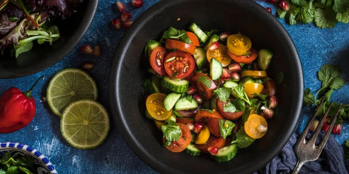 fresh salad with tomato, cucumber and herbs, flat lay from above the salad is in a black bowl and there are other bowls with ingredients around it the table is decorated with lime, chili and cilantro