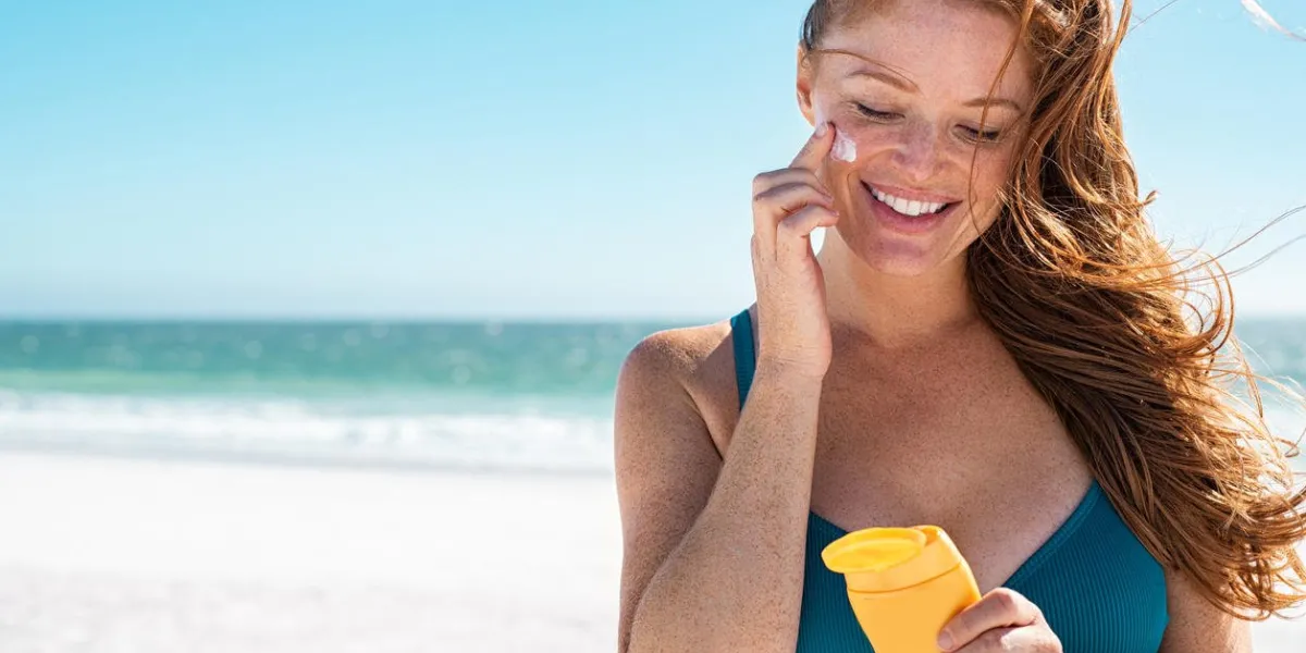 beautiful young woman in blue bikini at beach applying sunscreen on face for protection on a sunny day mature woman with freckles and red hair enjoying summer holiday while applying suntan lotion portrait of smiling lady with healthy skin