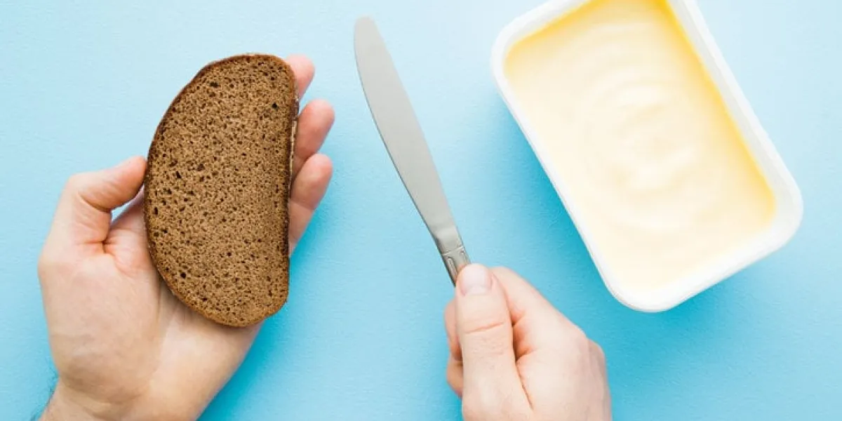 man's hands holding slice of brown bread and knife opened plastic pack of light yellow margarine on pastel blue desk preparing breakfast point of view shot closeup top view
