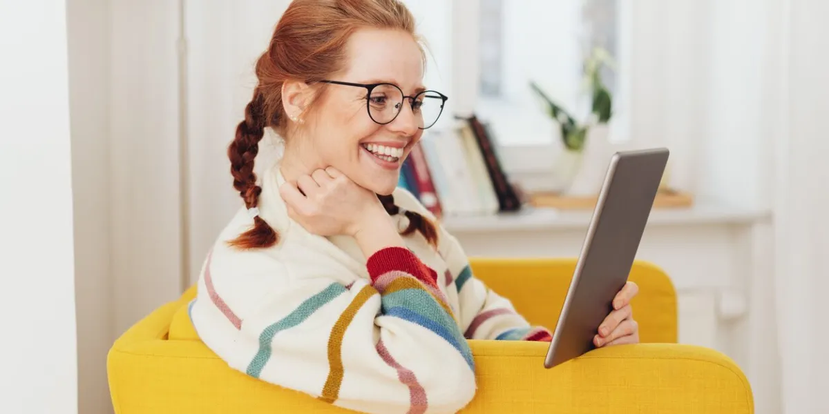 smiling happy young woman reading on a tablet as she relaxes in a colorful yellow armchair at home