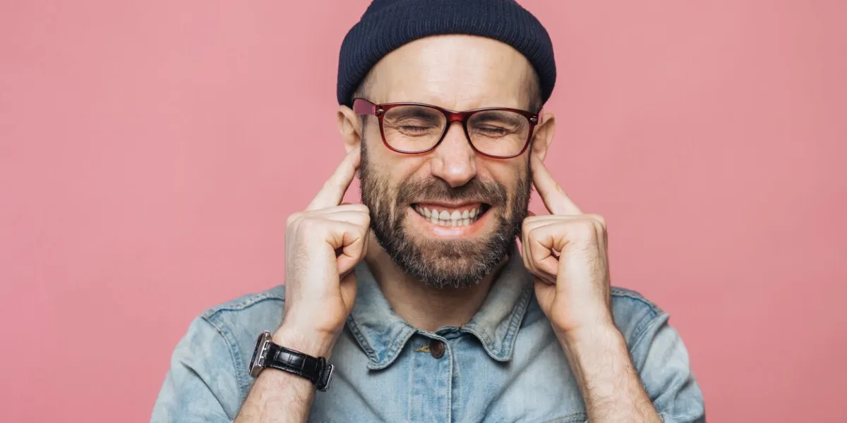 annoyed bearded male plugs ears as hears unpleasant sound, keeps eyes shut, being dissatisfied with something, isolated over pink background unshaven middle aged male ignores hearing someone