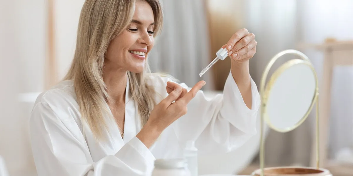 happy beautiful middle aged woman in bathrobe sitting in front of mirror, holding bottle with facial oil or serum, lady applying face care product cosmetics after morning shower, copy space