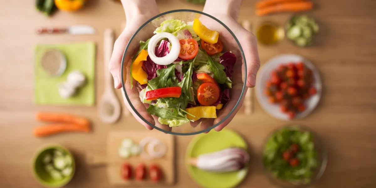 hands holding an healthy fresh vegetarian salad in a bowl, fresh raw vegetables on background, top view