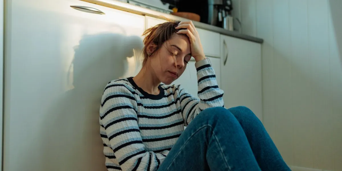 young woman sitting on the kitchen floor with her head in her hand, suffering from depression, loneliness, and mental health issues