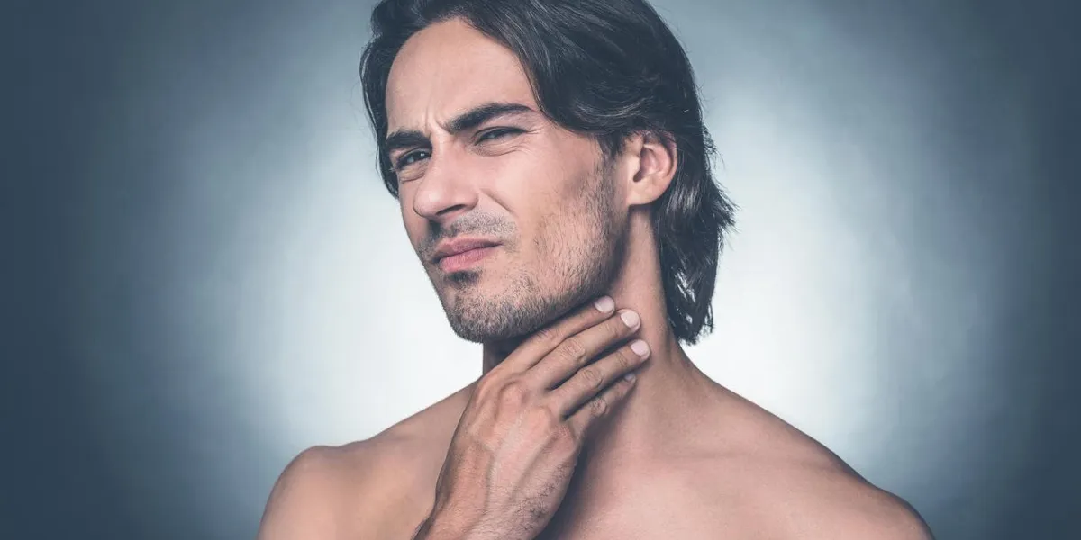 portrait de jeune homme torse nu, exprimant la négativité tout en touchant son cou et se tenant debout sur fond gris