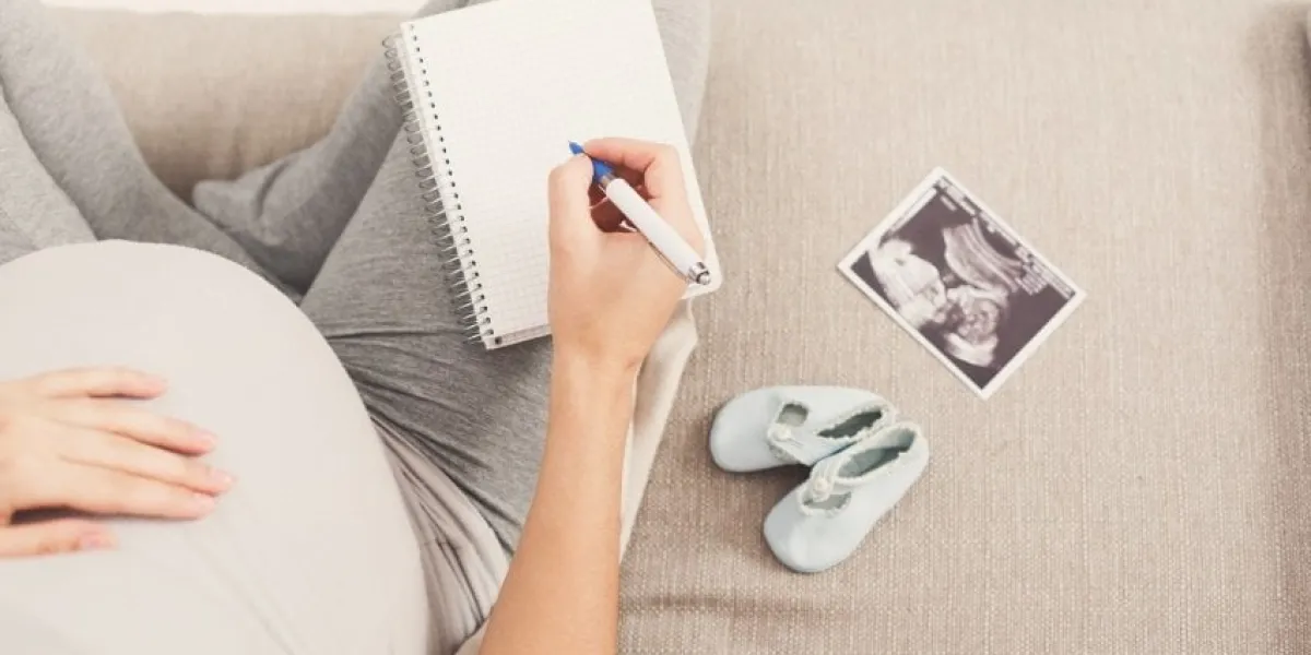 unrecognizable pregnant woman sitting on sofa with blank notebook, pen, tiny shoes and sonogram and writing names for her baby, making shopping list, top view, copy space