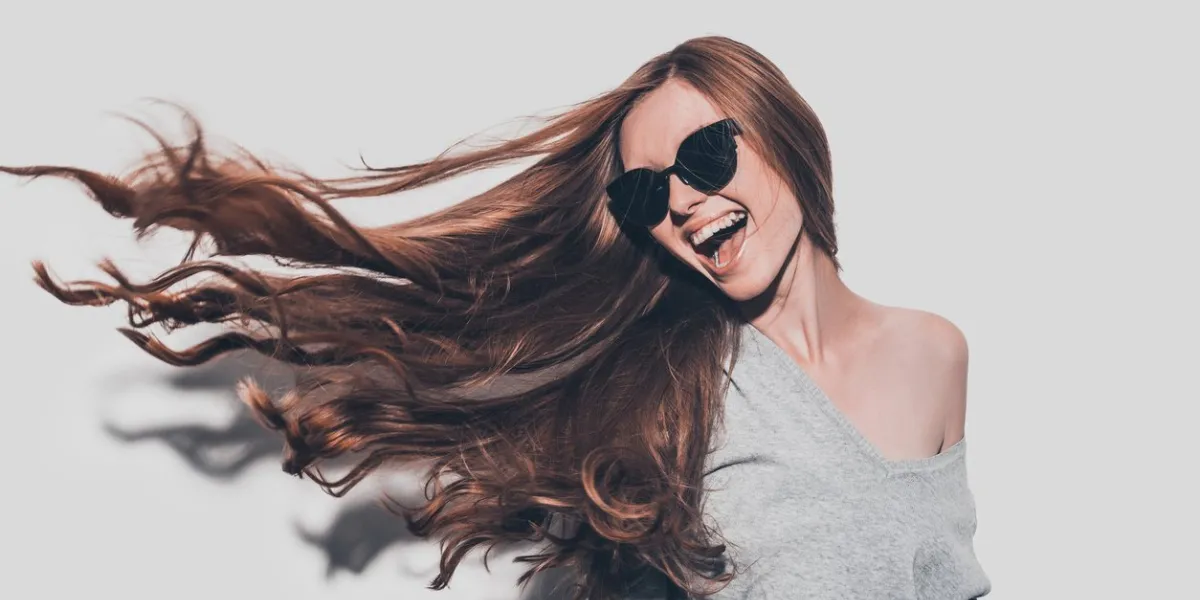 attractive young smiling woman in sunglasses and with tousled hair looking away while standing against grey background