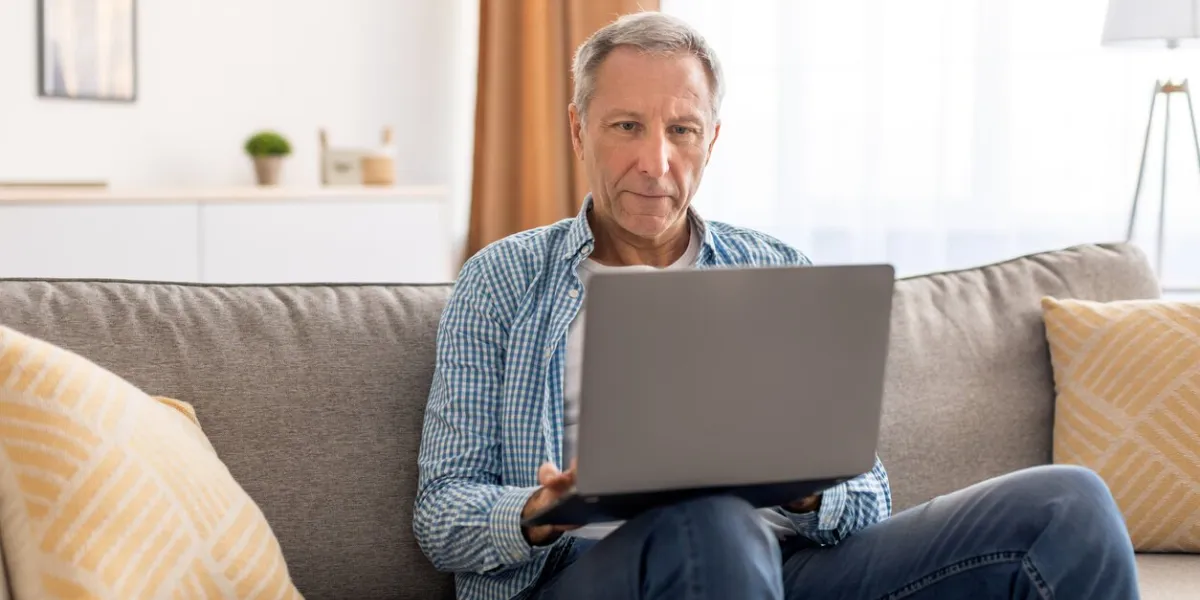 people and technology portrait of mature man using laptop sitting on the couch in living room confident senior male adult resting at home working on pc, looking at screen, typing on keyboard