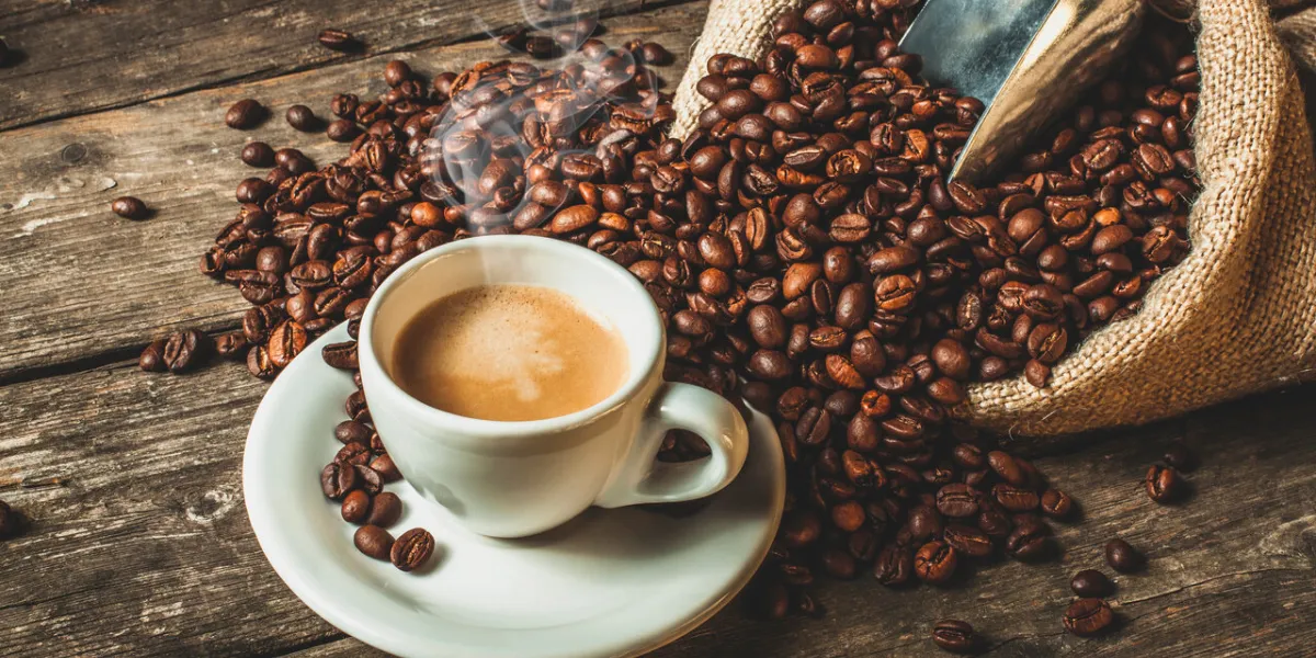 steaming coffee in traditional ceramic cup, jute sack with roasted beans on wooden table