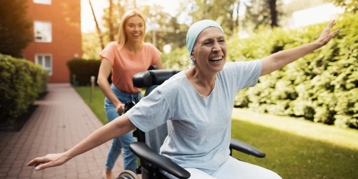 a woman with cancer is sitting in a wheelchair she walks on the street with her daughter and they fool around they are fun and they laugh they walk in the courtyard of the clinic