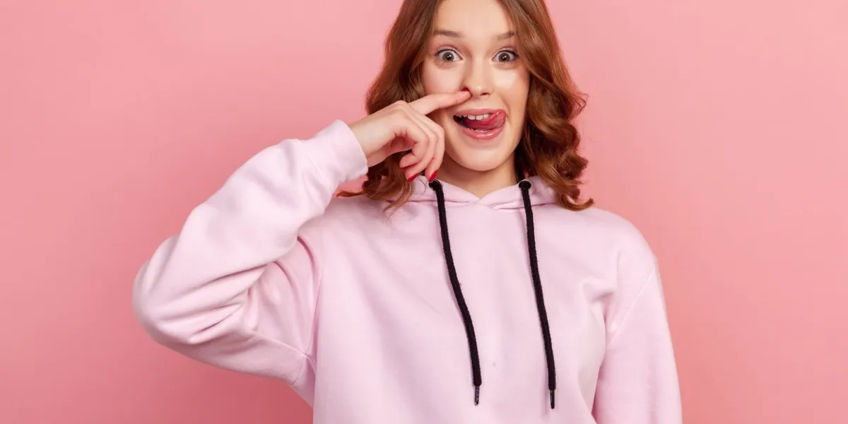 portrait of funny teen curly haired girl in hoodie drilling nose fooling around, showing tongue out and picking nose with comical silly expression indoor studio shot isolated on pink background