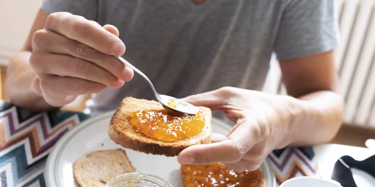 closeup of a young caucasian man, wearing a casual gray t-shirt, sitting at a set table, spreading some orange or peach jam on a toast