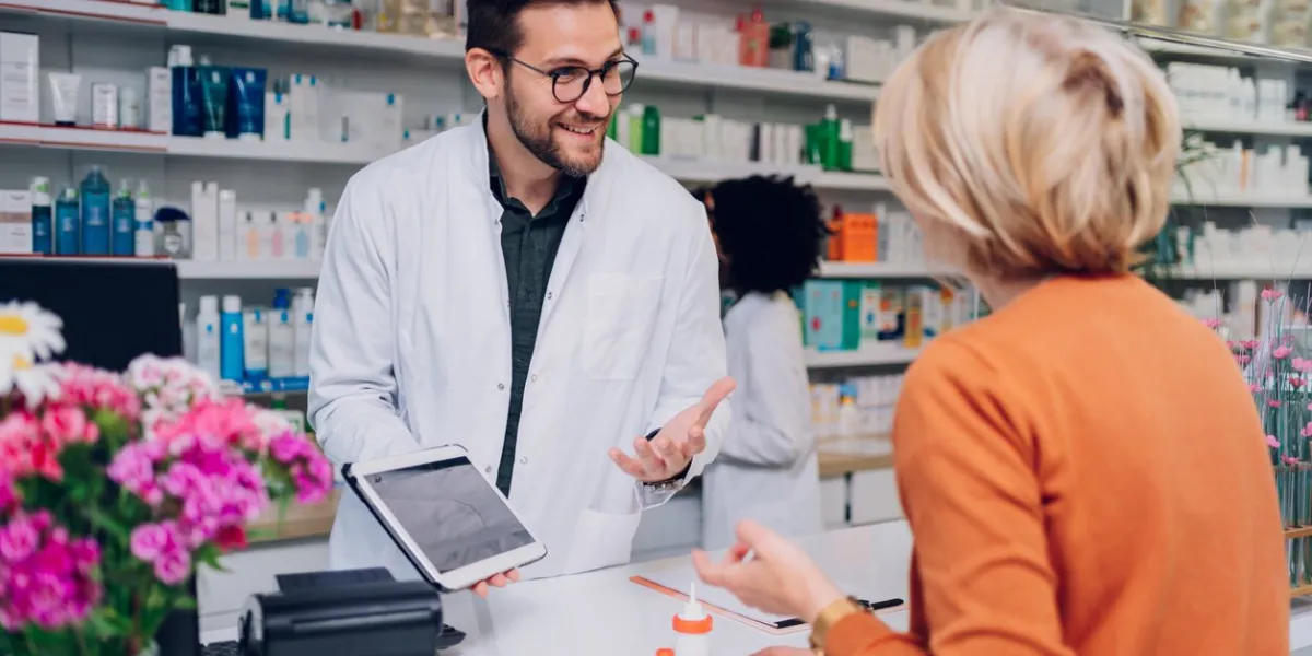 young male caucasian pharmacist showing tablet to senior female customer in a pharmacy with female pharmacist in the background shelves with health care products