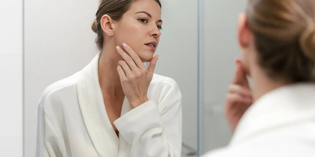 young and gorgeous woman in white bathrobe standing in bright light bathroom with mirror she looking at her reflection with calm face, holding hand on cheek, checking skin