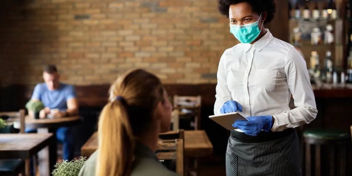 happy black waitress wearing protective face mask while taking order from customer on a touchpad in a cafe