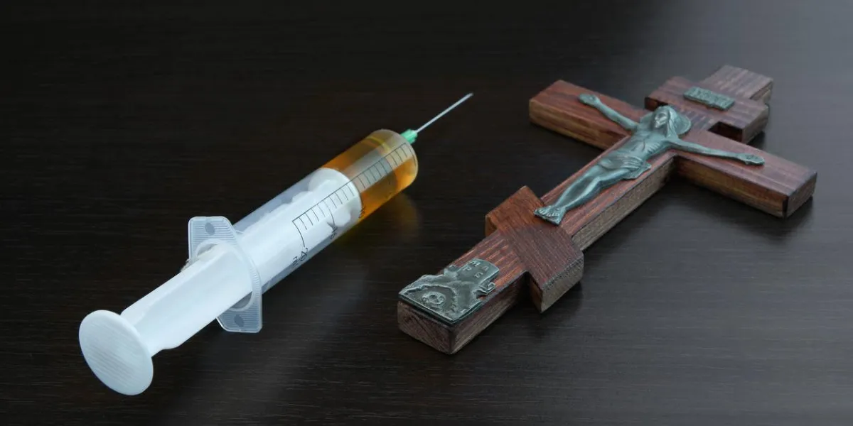 silver figurine of jesus christ crusified on red wood cross in spotlight and syringe with yellow liquid on black background, religion concept, closeup, top view