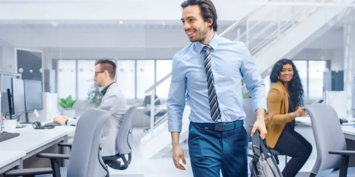 young happy businessman returns to work after vacation, walks into bright modern office, greets everybody, takes place at his desktop computer