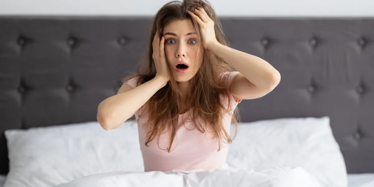 shocked millennial woman sitting on bed at home, holding her head in terror, being late for work terrified young caucasian lady oversleeping, having trouble waking up in morning