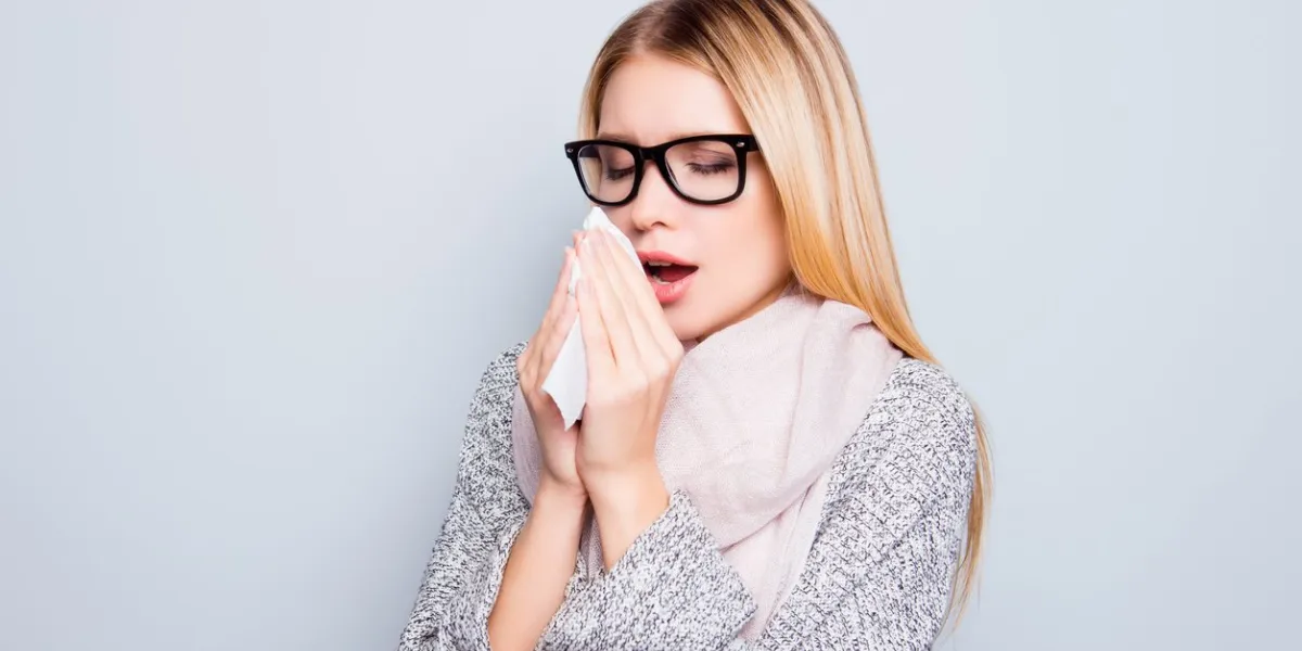 concept of catching a cold in autumn close up portrait of young woman suffering from a cold she is sneezing and using a paper tissue she is wearing warm cardigan and scarf isolated on background