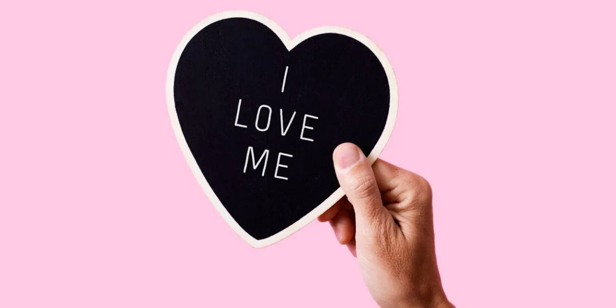 closeup of a man holding a black heart-shaped sign with the text i love me written in it, on a pink background