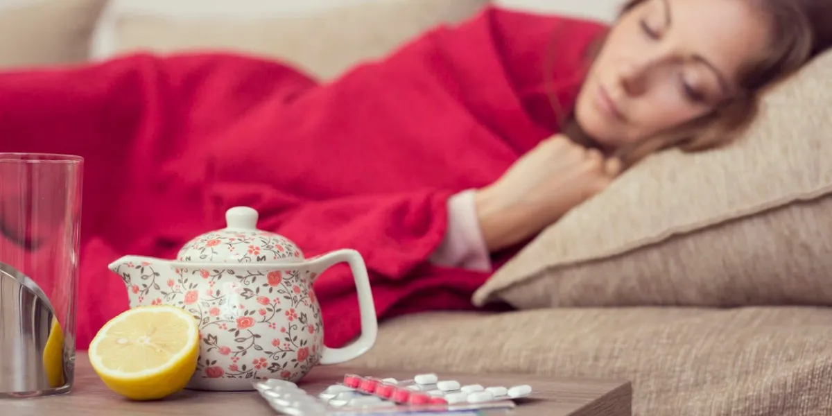 sick woman covered with a blanket lying in bed with high fever and a flu, resting teapot, pills and lemon on the table, focus on the teapot