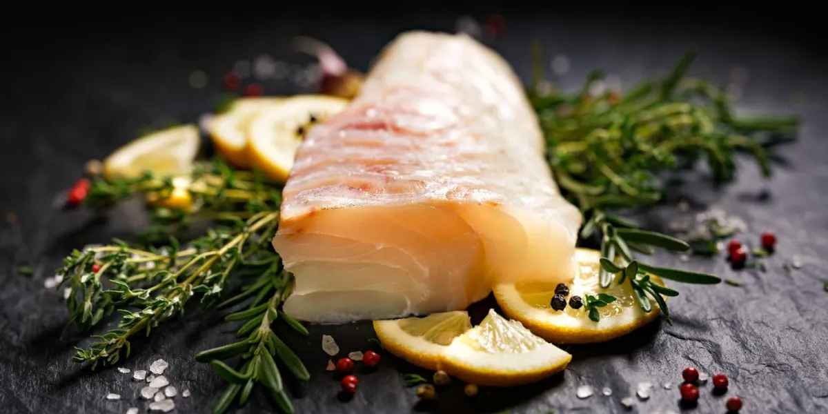 fresh raw cod fillet with addition of herbs and lemon slices on black stone background