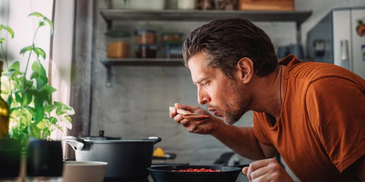 close up shot of a handsome young happy caucasian man tasting sauce with a mixing spoon with his eyes closed over a frying pan in a kitchen