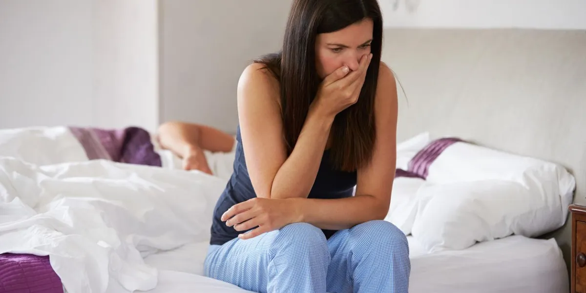 woman sitting on bed feeling sick, while man sleeps