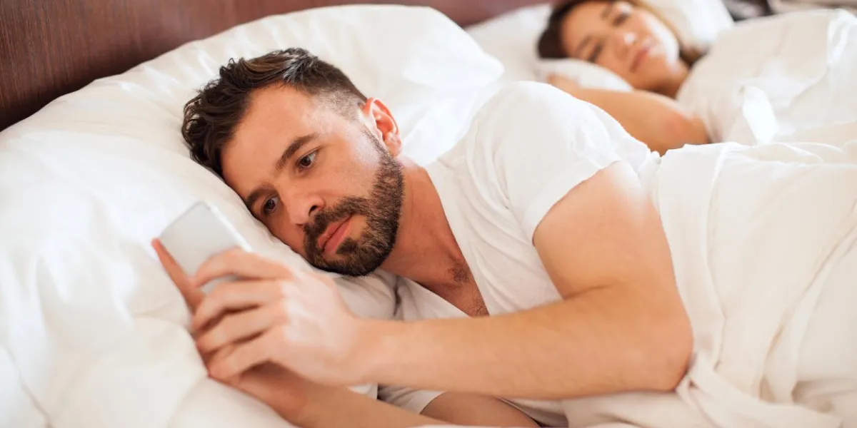 portrait d'un beau jeune homme avec une barbe vérifiant son smartphone et envoyer des sms à son amant pendant que son partenaire dort