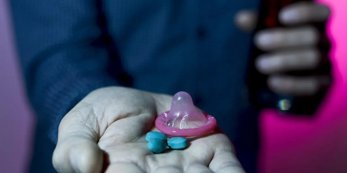 closeup of a young caucasian man with some blue pills and a condom in one hand and a glass with a mixed drink in the other hand, in a night club