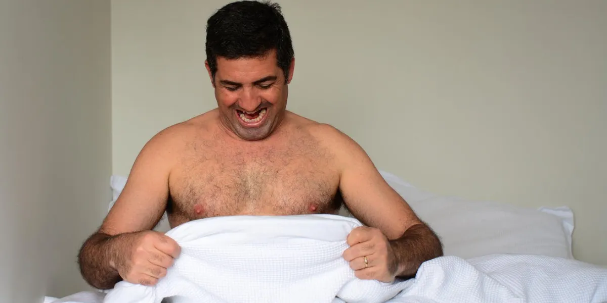 happy man in his forties (40s) in bed looking down at his penis under white covers sheet in bedroom concept photo of male sexuality and man sex and relationship problem concept