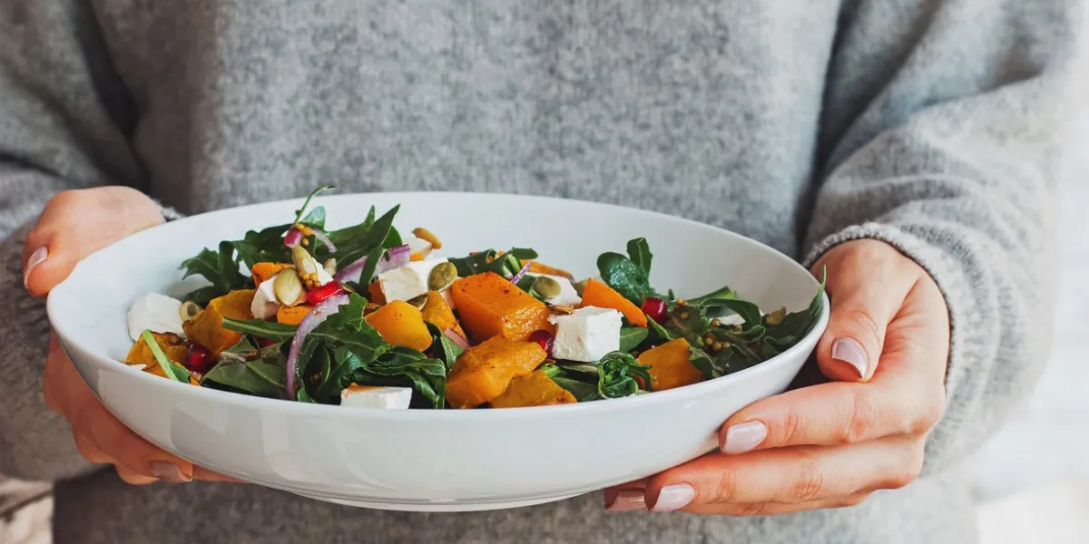 woman holding a bowl of salad with pumpkin, arugula, feta cheese and pomegranate seeds cozy comfort food concept