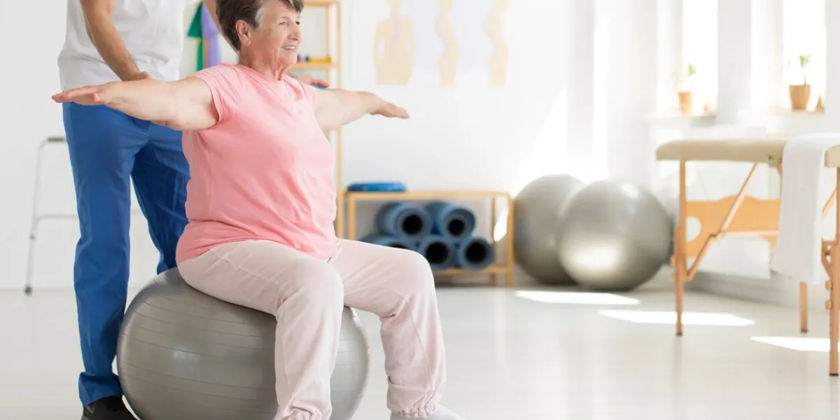 elderly senior ward trying to maintain balance while sitting on a grey fit ball while being supported by physiatrist