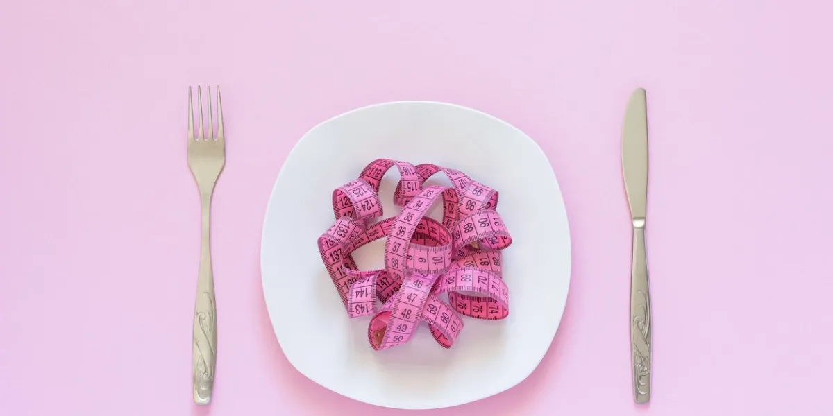 pink measuring tape lying on plate in the form of spaghetti, knife and fork on pink background diet or anorexia concept top view