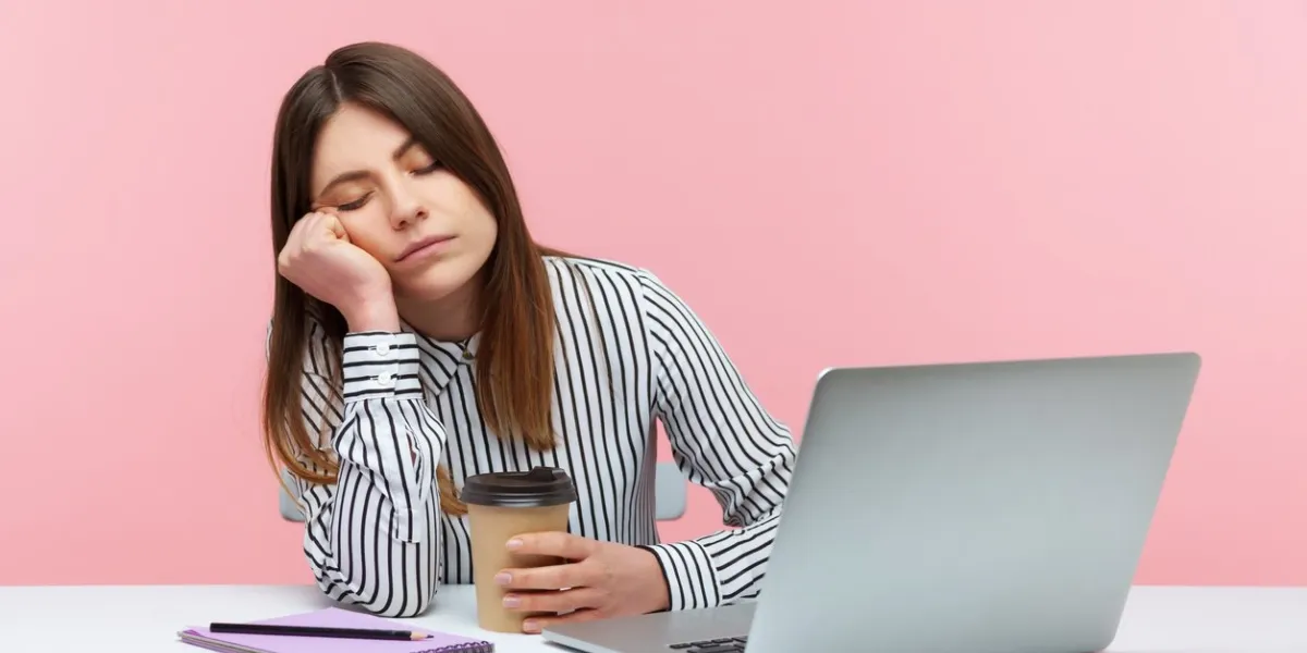 sleepy inefficient woman office worker napping leaning head on hand and holding coffee cup sitting at workplace with laptop, physical exhaustion indoor studio shot isolated on pink background