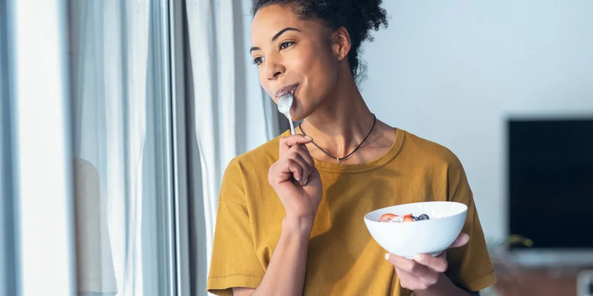 shot of beautiful mature woman eating cereals and fruits while standing next to the window at home
