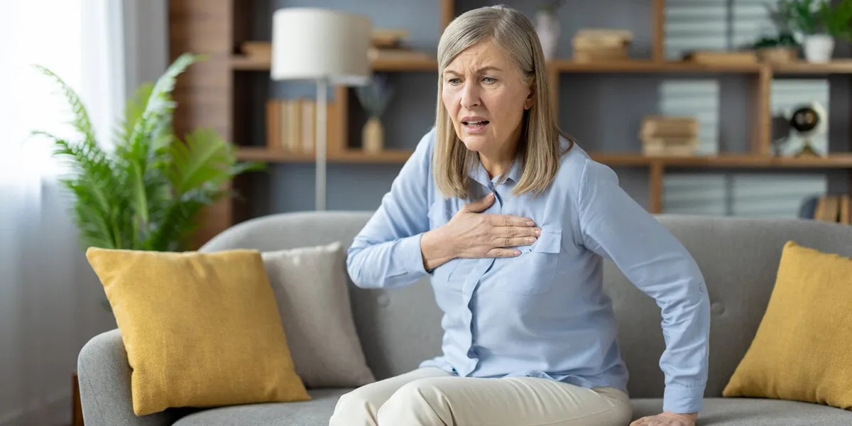 elderly female grabbing chest with palm while sitting on pillowed couch with shocked expression ill woman feeling sharp cardiac pain and risk of infarction while sitting alone in modern flat