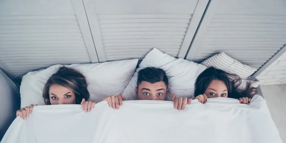 top view of funny three partners hiding behind blanket, peeking over sheets and looking at camera while lying in bed at home, weekend, rest, relax, someone catches them