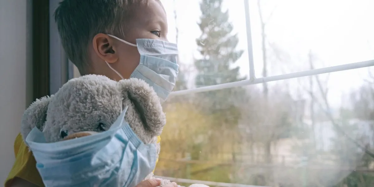 sad illness child on home quarantine boy and his teddy bear both in protective medical masks sits on windowsill and looks out window virus protection, coronavirus pandemic, prevention epidemic