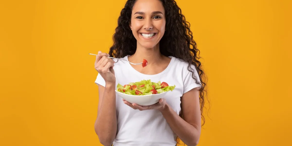 healthy meal portrait of happy latin casual woman eating tasty fresh vegetable salad, holding plate bowl and fork looking at camera satisfied millennial lady isolated on yellow orange background
